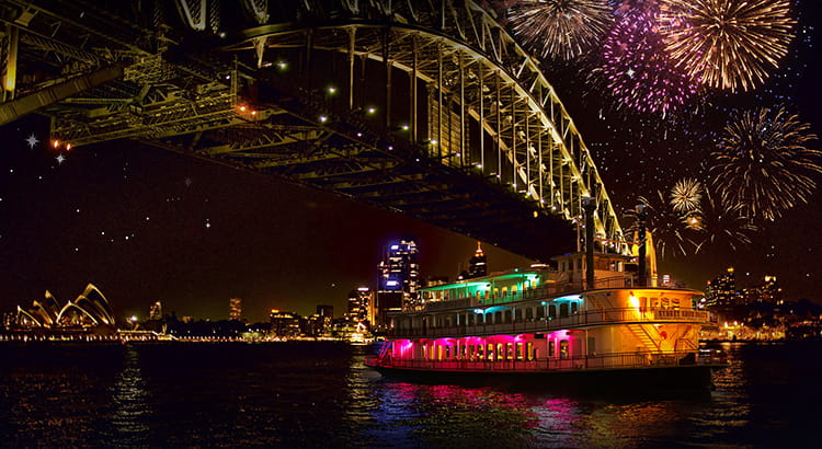 Sydney Showboats cruises past the Harbour Bridge illuminated by the NYE fireworks in the night sky.